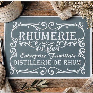 Peut inclure: Un pochoir rectangulaire avec un fond gris foncé, du texte blanc et des bordures à volutes. Le texte indique RHUMERIE, Entreprise Familiale et DISTILLERIE DE RHUM dans une police décorative avec un motif central de fleur de lys.