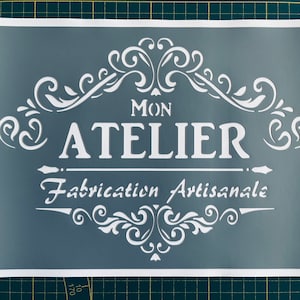 Puede incluir: Una plantilla gris con letras blancas que dice "Mon Atelier Fabrication Artisanale" con adornos decorativos.