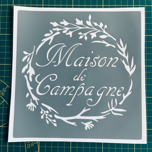 Op de afbeelding: Een grijze sjabloon met de woorden "Maison de Campagne" in een decoratief lettertype, omringd door een witte bloemenkrans. De sjabloon staat op een witte achtergrond.