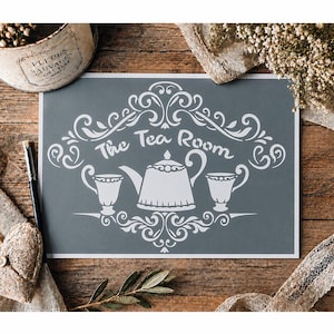 Peut inclure: Une feuille de pochoir rectangulaire avec un fond gris foncé et un motif graphique blanc. Le design comprend le texte The Tea Room, une théière centrale et deux tasses, le tout entouré de volutes et d'ornements blancs complexes.