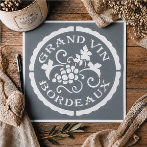 Op de afbeelding: Een vierkant sjabloon met een cirkelvormig ontwerp en een geschulpte rand. De witte afbeelding bevat in het midden een tros druiven met bladeren, omringd door de tekst GRAND VIN BORDEAUX in een vetgedrukt schreeflettertype op een grijze achtergrond.