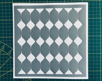 Op de afbeelding: Een grijze en witte stencil met een herhalend patroon van afgeronde ruitvormen. De stencil is gemaakt van een dun, flexibel materiaal en is ontworpen voor gebruik met verf of andere media.
