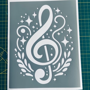 Peut inclure: Un pochoir en vinyle blanc d'une clé de sol avec un motif floral décoratif et des étoiles. Le pochoir est sur un fond gris.