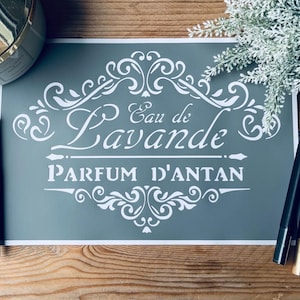Peut inclure: Un panneau gris avec un texte blanc orné qui dit "Eau de Lavande Parfum d'Antan". Le panneau est posé sur une surface en bois, avec un pinceau de maquillage, une bougie et des stylos à proximité. Une petite plante est dans le coin supérieur droit.