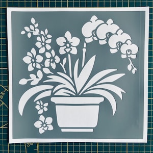 Op de afbeelding: Witte sjabloon van een orchideeënplant in een pot op een grijze achtergrond. De sjabloon is ontworpen voor knutselen en decoreren.