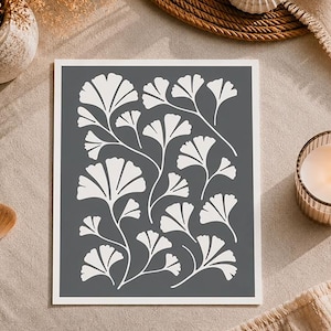 Könnte beinhalten: Gerahmter Druck mit einem weißen Ginkgo-Blattmuster auf dunklem Grauton. Die botanische Illustration hat einen weißen Rand. Der Druck wird auf einer neutralen Oberfläche mit einer Kerze und dekorativen Elementen im Hintergrund präsentiert.