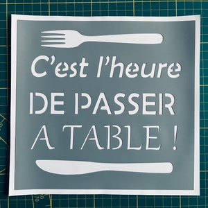 May include: A square sign with a grey background and white text and cutlery. The text reads "C'est l'heure DE PASSER A TABLE!" A white fork is at the top and a white knife at the bottom.