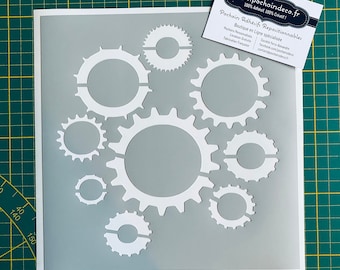 May include: A white stencil with a design of multiple interlocking gears. The stencil is on a gray background.