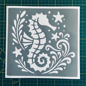 May include: A gray stencil with a white seahorse design. The seahorse is surrounded by stylized waves and starfish.