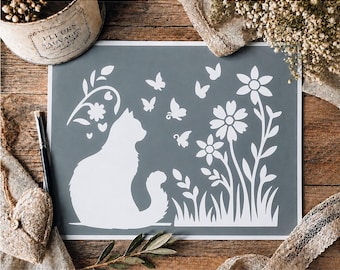 Könnte beinhalten: Eine rechteckige Schablonenfolie mit einem weißen Silhouettenmotiv auf grauem Hintergrund. Das Design zeigt eine sitzende Katze, die einer Ansammlung von Blumen, Gras und mehreren Schmetterlingen gegenübersteht. Ein dekoratives Rankenelement befindet sich über der Katze.