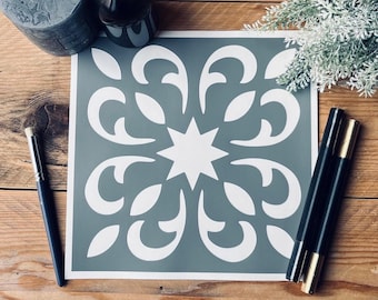 Op de afbeelding: Een vierkante sjabloon met een symmetrisch bloemmotief in wit op een grijze achtergrond. Een penseel en twee zwarte stiften met gouden accenten liggen naast de sjabloon. De items liggen op een houten oppervlak.