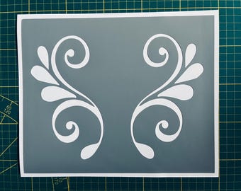 Op de afbeelding: Een grijze stencil met een wit ontwerp van twee symmetrische swirls met bladeren. De stencil ligt op een groene snijmat.
