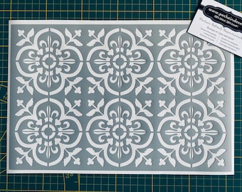 May include: A white stencil with a repeating pattern of a stylized floral design on a gray background. The stencil is designed for use with paint or other mediums to create a decorative tile effect.