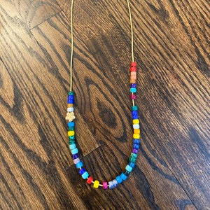 Op de afbeelding: Een kleurrijke kralenketting met een regenboog aan kleuren, waaronder rood, oranje, geel, groen, blauw, indigo en violet. De ketting is gemaakt van kleine, vierkante kralen en heeft een gouden bedel in het midden.