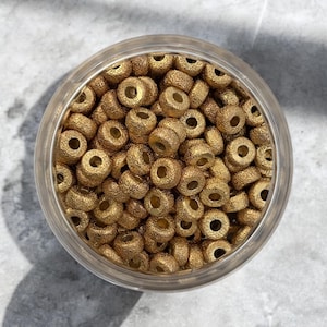 Op de afbeelding: Een close-up van een glazen pot gevuld met gouden kralen. De kralen zijn rond en hebben een getextureerd oppervlak.