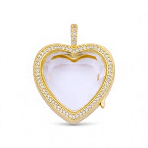 May include: A gold-colored heart-shaped pendant with a clear center. The pendant is outlined with small, clear gemstones. The bail is also adorned with gemstones. The pendant is designed to hold a small photo or memento.