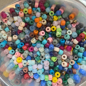 Op de afbeelding: Een kom gevuld met kleurrijke, ronde en vierkante kralen. De kralen zijn gemaakt van verschillende materialen, waaronder glas, steen en metaal. De kralen zijn in verschillende kleuren, waaronder blauw, groen, rood, geel en paars.