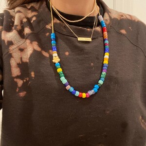 Op de afbeelding: Een kleurrijke kralenketting met een kleine bloembedel. De ketting is gelaagd met twee gouden kettingen, een met een klein rechthoekig naamplaatje met de tekst "NATH + SYDNEY".