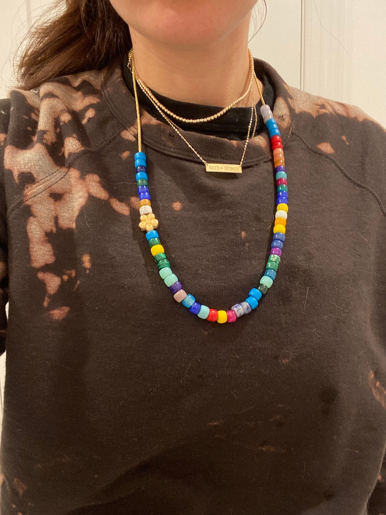 Op de afbeelding: Een kleurrijke kralenketting met een kleine bloembedel. De ketting is gelaagd met twee gouden kettingen, een met een klein rechthoekig naamplaatje met de tekst "NATH + SYDNEY".
