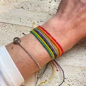 Puede incluir: Una muñeca con un brazalete de cadena plateada y siete pulseras de cuentas de colores en tonos de rojo, naranja, amarillo, verde, azul, morado y rosa.
