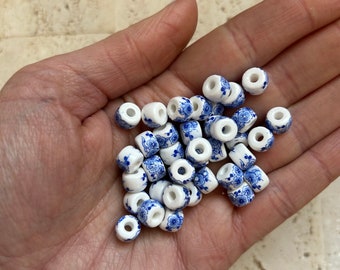 May include: A collection of white ceramic beads with a blue floral pattern. The beads are cylindrical with a hole through the center, suitable for jewelry making or crafts. The beads are small and delicate.