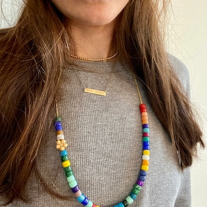Op de afbeelding: Een gouden ketting met een kleurrijke kralenketting en een klein gouden bloemachtig sieraad. De ketting heeft ook een dunne gouden ketting met een staafvormig sieraad met de tekst "with + sydnee".