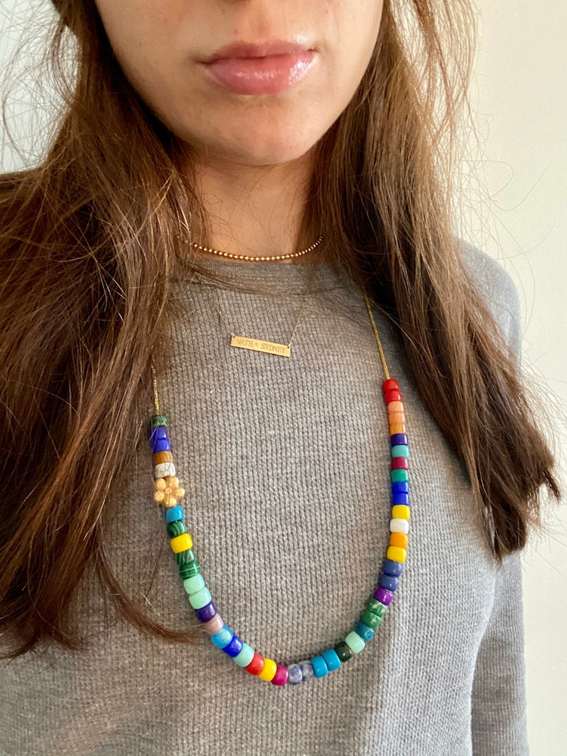Op de afbeelding: Een gouden ketting met een kleurrijke kralenketting en een klein gouden bloemachtig sieraad. De ketting heeft ook een dunne gouden ketting met een staafvormig sieraad met de tekst "with + sydnee".