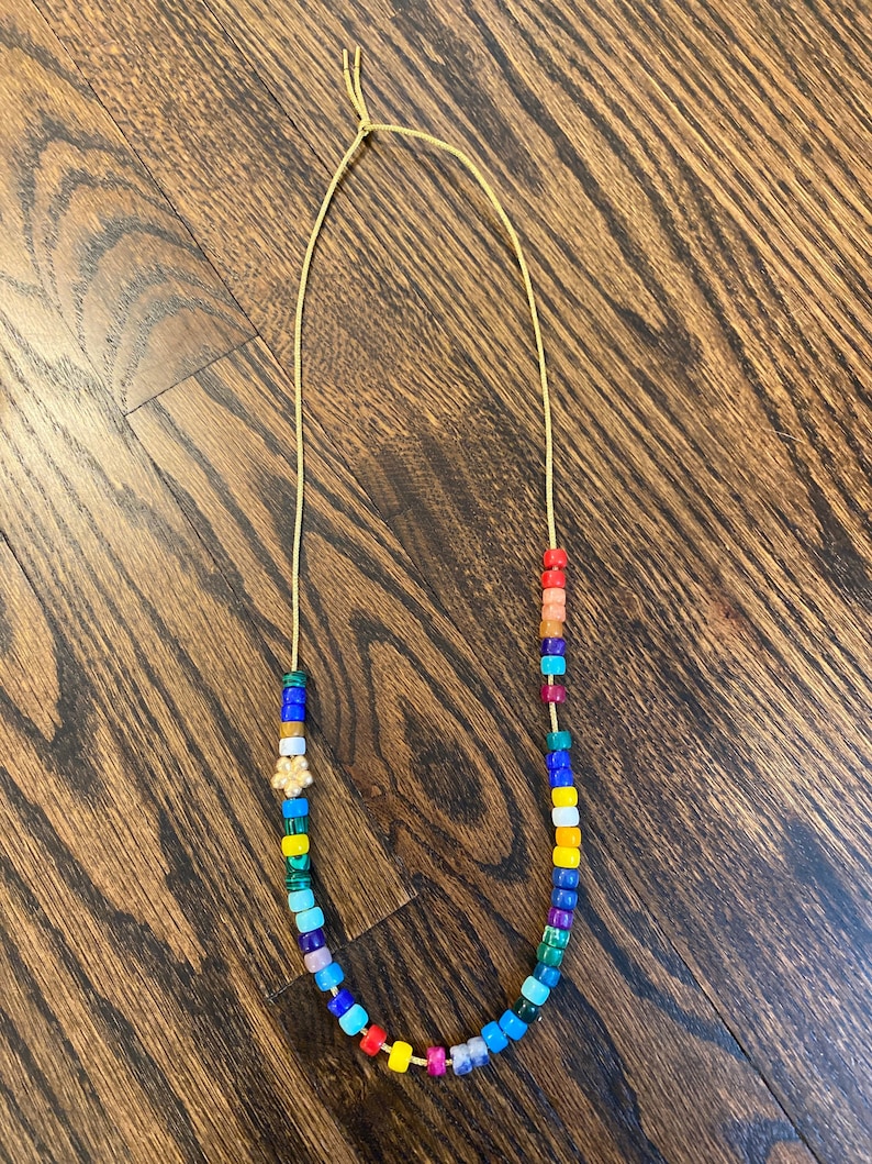 Op de afbeelding: Een kleurrijke kralenketting met een regenboog aan kleuren, waaronder rood, oranje, geel, groen, blauw, indigo en violet. De ketting is gemaakt van kleine, vierkante kralen en heeft een gouden bedel in het midden.