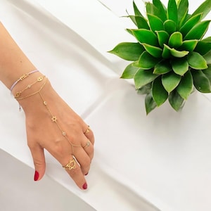 May include: A gold chain bracelet with a Star of David charm and a small eye charm. The bracelet is layered with a thin gold chain bracelet and a purple string bracelet with the word "mila" on it. The bracelet is worn on a hand with gold rings, including a Star of David ring.