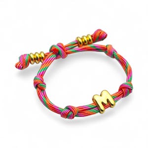 Peut inclure: Un bracelet réglable coloré avec un charm en forme de lettre "M" de couleur or. Le bracelet est composé d'un cordon tissé dans les tons de rouge, orange, vert et rose. Des perles dorées ornent la fermeture réglable.