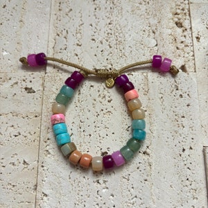 Op de afbeelding: Een kleurrijke kralen armband met verschillende stenen, waaronder turquoise, roze en groen. De armband is vastgemaakt met een bruin koord en heeft een gouden sluiting.