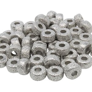 Op de afbeelding: Een stapel zilverkleurige metalen kralen met een getextureerd oppervlak. De kralen zijn rond met een gat in het midden.