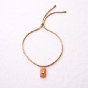 May include: A necklace with a peach-colored rectangular pendant and a small, square, gold-colored accent. The necklace has a gold-colored cord and adjustable closure. The jewelry is displayed on a white background.