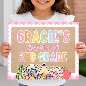 May include: A rectangular sign with a pink scalloped border and a kraft paper background. The text reads "GRACIE'S first day of 2ND GRADE" in colorful letters. Below the text are illustrations of a pear, a pencil, and an apple.