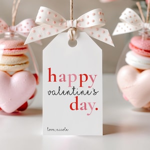 May include: A white gift tag with the text "happy valentine's day" in red and pink. The tag is tied with a dotted pink bow. In the background are glass ornaments filled with macarons and heart-shaped candies.