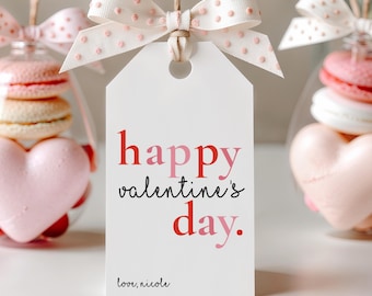 May include: A white gift tag with the text "happy valentine's day" in red and pink. The tag is tied with a dotted pink bow. A clear glass ornament with pink macarons and a heart-shaped candy is visible.