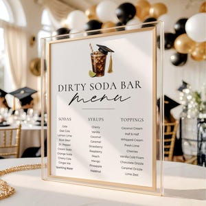 May include: A clear acrylic sign with a gold frame displays a "Dirty Soda Bar Menu." The menu lists sodas, syrups, and toppings. A graduation cap and a drink illustration are at the top. The background includes balloons and tables.