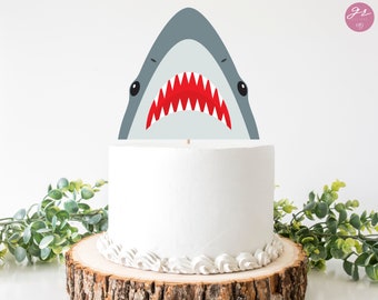 Puede incluir: Un pastel blanco con un adorno de tiburón gris con dientes rojos. El pastel está sobre un soporte para pasteles de madera.