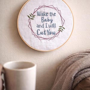 Könnte beinhalten: Gestickte Stickrahmenkunst mit dem Text "Wake the Baby and I will Cut You" in blauem Faden. Das Design umfasst einen geometrischen Rahmen und grüne Blattakzente. Ein Holzreifen hält den Stoff.