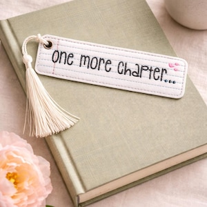Puede incluir: Un marcapáginas blanco con el texto "one more chapter..." en escritura negra, con una borla color crema. El marcapáginas está sobre un libro verde salvia, con una flor rosa y una taza de café en el fondo.