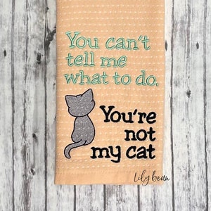 以下が含まれることがあります： 桃色のキッチンクロスに、灰色の猫のイラストと「You can't tell me what to do. You're not my cat.」というテキストが刺繍されています。テキストは緑と黒で刺繍されています。クロスには白い水玉模様があります。
