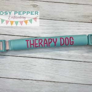Puede incluir: Una correa de perro de tela azul turquesa con una hebilla de plástico y las palabras "Therapy Dog" bordadas en rosa.