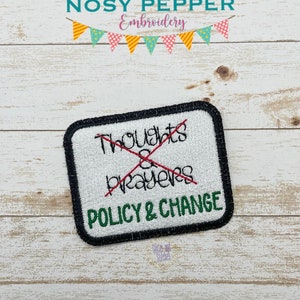 Puede incluir: Un parche blanco bordado con costuras negras alrededor de los bordes. El parche tiene una línea roja dibujada a través de las palabras "Thoughts & Prayers" y las palabras "Policy & Change" escritas debajo en verde.