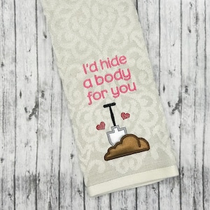 May include: White kitchen towel with pink embroidery that reads "I'd hide a body for you" with a pink shovel and two pink hearts above a brown mound of dirt.