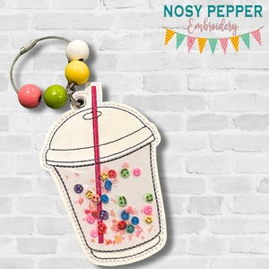 May include: A bubble tea-shaped keychain in white and clear, featuring a pink straw and colourful smiley face confetti. It has a beaded accent with pink, green, yellow, and white beads. The text "AUGUST 2025 MEGA BUNDLE" is displayed at the bottom of the image.