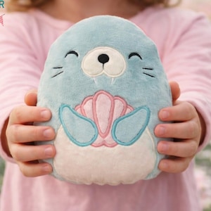 May include: A plush seal toy in shades of blue, pink, and white. The seal has a smiling face with black eyes and whiskers. A pink shell design is on the seal's belly. The toy is held in front of a blurred floral background. The text "Nosy Pepper Embroidery" is in the top left corner.