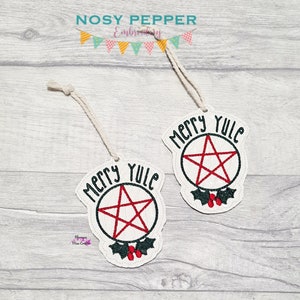 May include: Two white felt Christmas ornaments with the text "Merry Yule" and a red pentagram inside a black circle. Each ornament has a green holly sprig with red berries.