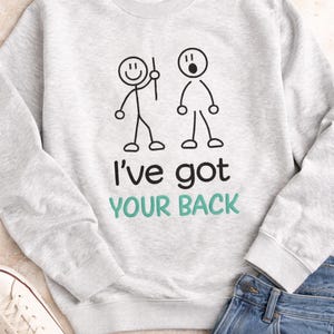 Può includere: Una felpa grigio chiaro con la scritta "I've got your back" in nero e verde acqua. La felpa presenta una grafica con omini stilizzati. Nell'immagine sono presenti anche jeans blu e scarpe da ginnastica bianche.