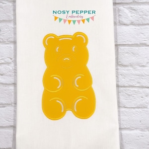 May include: A white towel with a large, embroidered yellow gummy bear design. The bear has a smiling face and silver accents. The text "Nosy Pepper Embroidery" is at the top, with a banner of colorful flags.