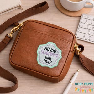 Può includere: Borsa a tracolla in pelle marrone con cerniera e cinturino dorati. La borsa presenta una toppa con la scritta "Maybe Swearing Will Help" in un design azzurro e bianco. È visibile una penna bianca.